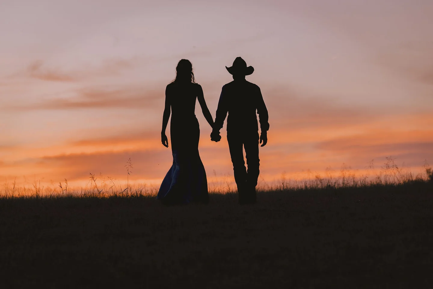 Silhouettes of the couple holding hands against an orange and pink country sunset.